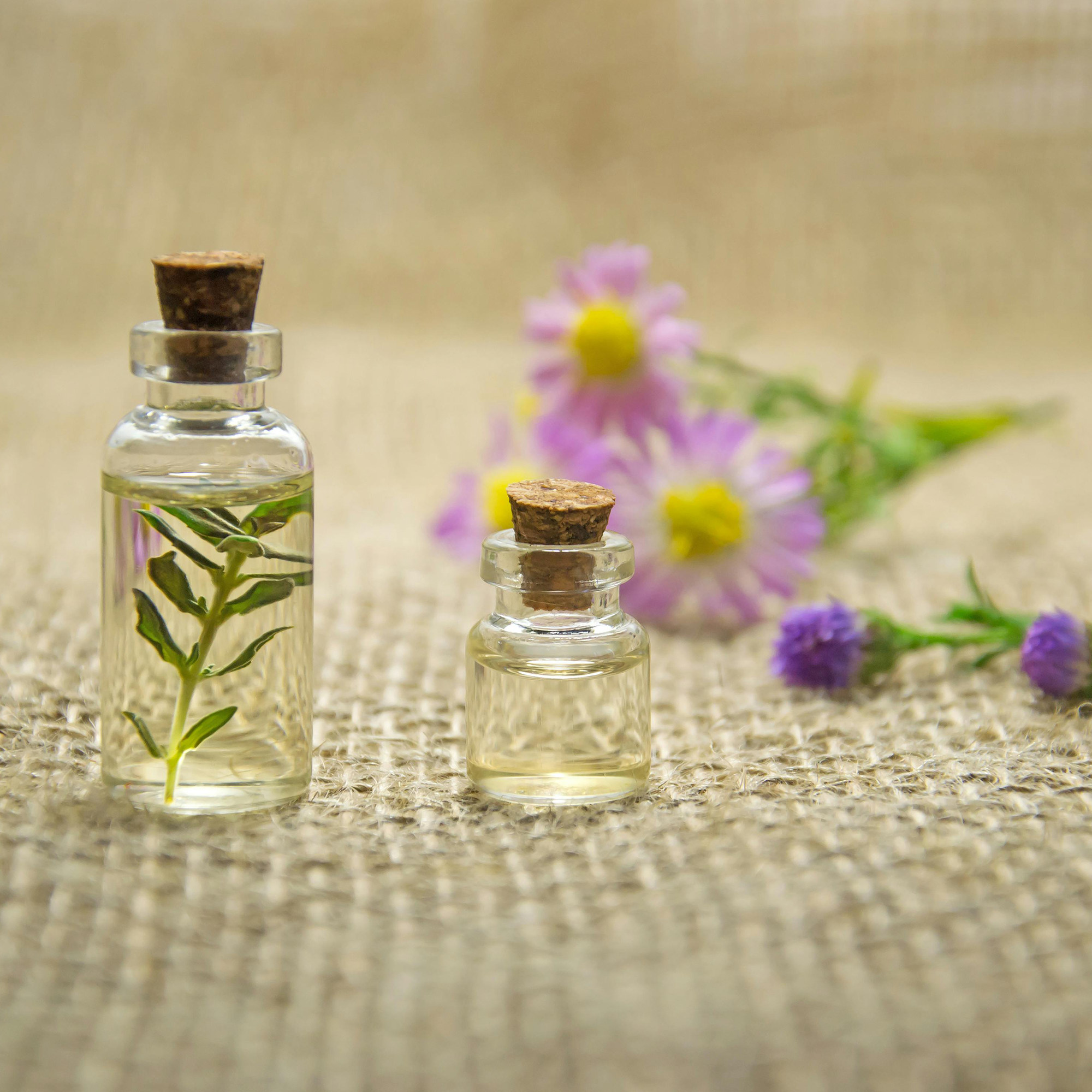 Two small glass bottles with cork lids containing essential oils, one with leaves inside, next to purple and yellow flowers on a burlap surface