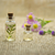 Two small glass bottles with cork lids containing essential oils, one with leaves inside, next to purple and yellow flowers on a burlap surface